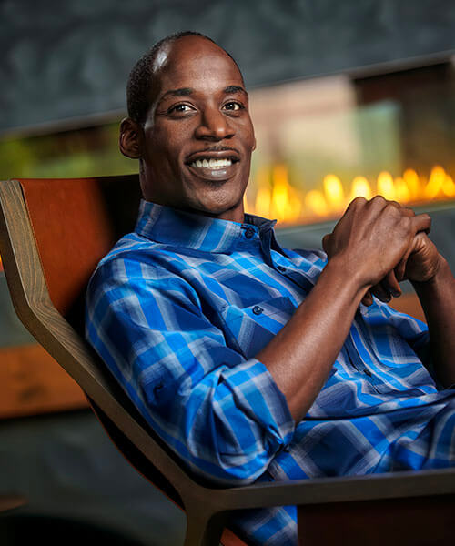 A man is smiling while seated in a chair, wearing a blue plaid shirt, in a cozy indoor setting.