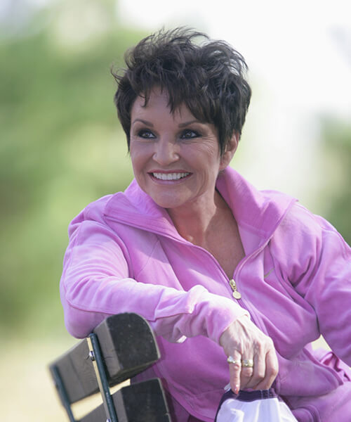 A woman in a pink tracksuit smiles while sitting on a bench outdoors.