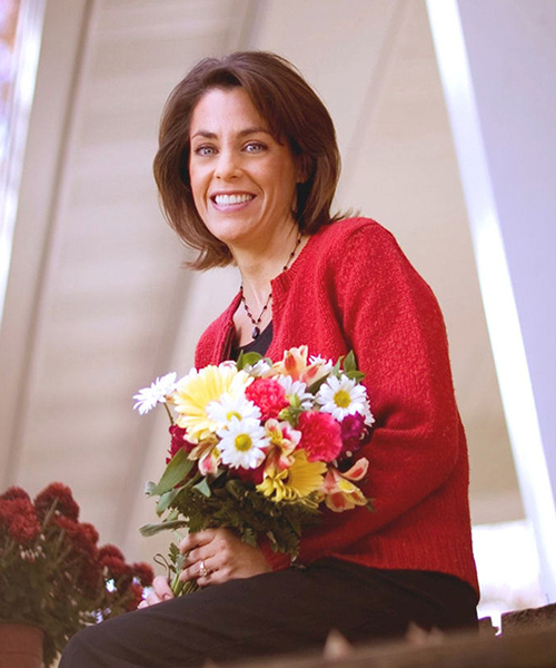 A person holding a bouquet of flowers, smiling outdoors highlighting her natural smile.